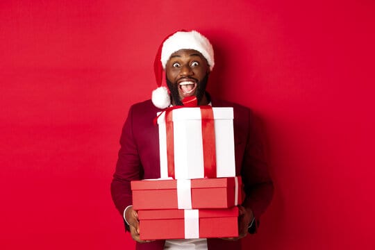 Christmas, New Year and shopping concept. Happy Black man in santa hat and blazer holding xmas prese