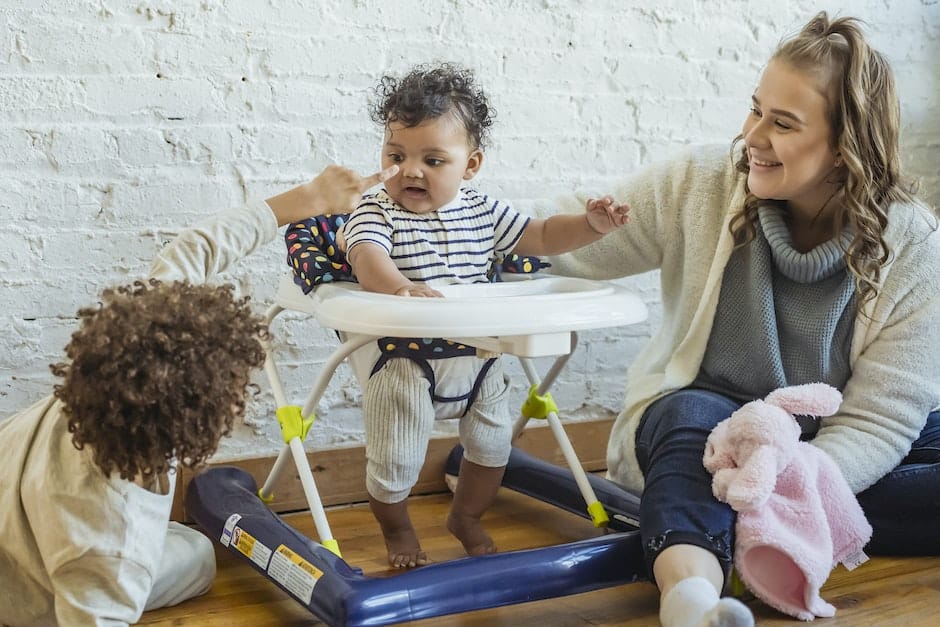 Happy mother with toy playing with black toddler in baby walker and boy while sitting on floor near 