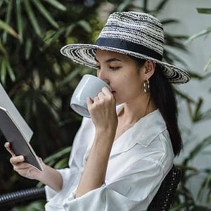 Young ethnic woman reading notebook while drinking beverage on chair