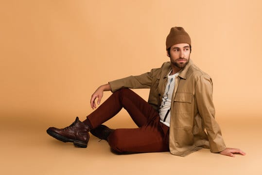 trendy man in stylish autumn outfit looking away while sitting on beige